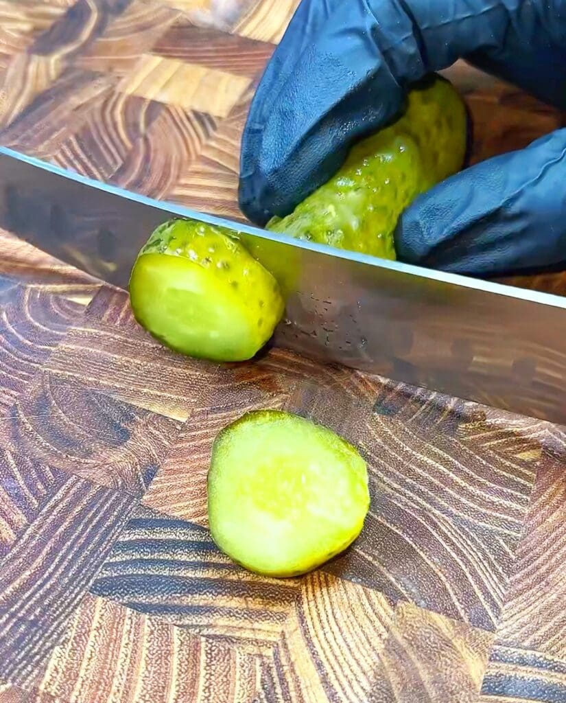 A gloved hand holds a pickle as a knife slices off a piece on a wooden cutting board.