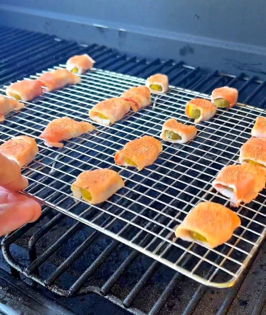 Bacon-wrapped pickle bites on a grill, with a hand holding the metal rack.