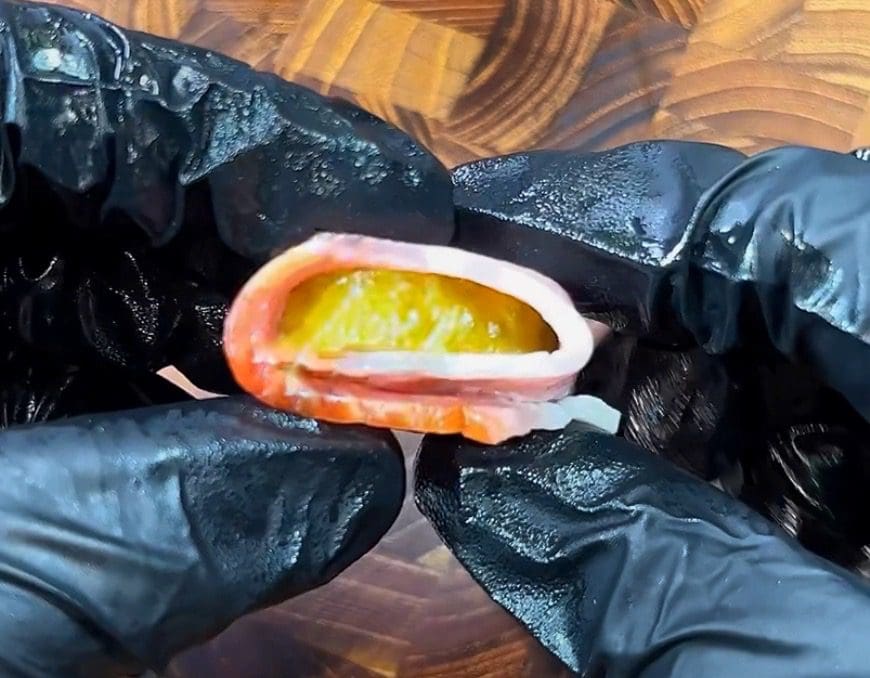 A person wearing black gloves holds a piece of food with layers, including a strip of fish wrapped around something orange, possibly fruit, against a wooden background.