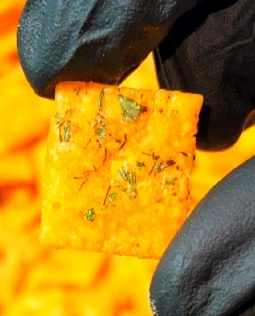 A person wearing black gloves holds a square, cheese-flavored cracker with visible herbs called Cool Ranch Cheez Its. The background reveals a tempting pile of similar snacks.