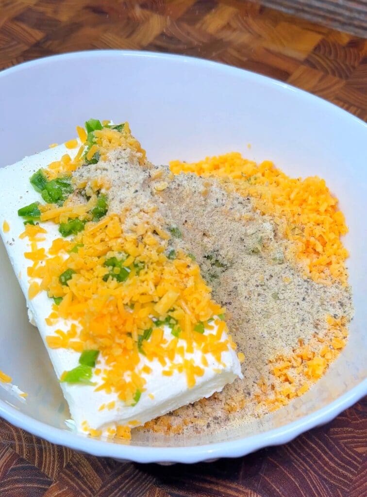 A white bowl contains a block of cream cheese topped with shredded cheddar, chopped green onions, and seasoning. for making a jalapeno cheese ball appetizer.
