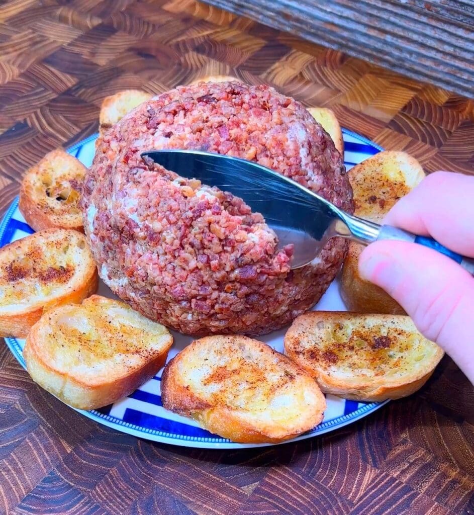 A hand holding a cheese knife slices into a crispy bacon covered jalapeno cheese ball with cheddar and cream cheese on a blue and white plate surrounded by toasted crostini bread rounds surrounded by toasted bread slices on a blue and white striped plate, set on a wooden surface.