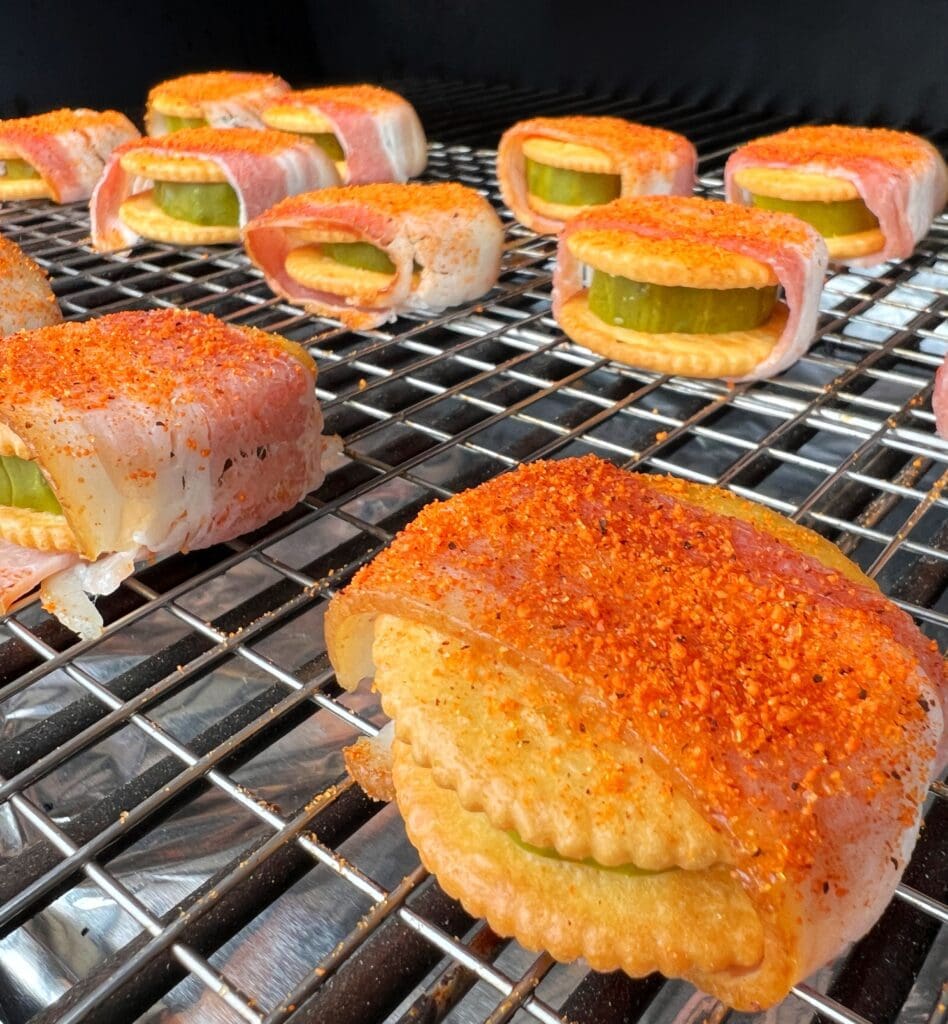 ritz crackers with pickles on the inside are wrapped with bacon, covered with seasoning, then  placed on the smoker.