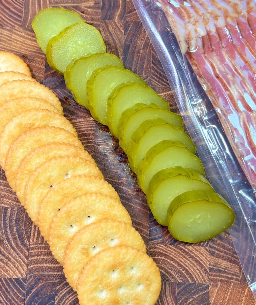 crackers, pickle, bacon on a wooden cutting board to make bacon wrapped crackers filled with dill pickles.
