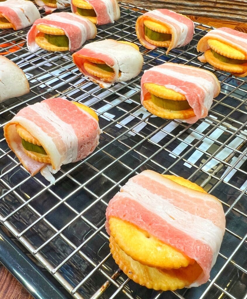 Crackers topped with pickles and wrapped in bacon are arranged on a wire rack over a baking sheet, ready to be cooked.