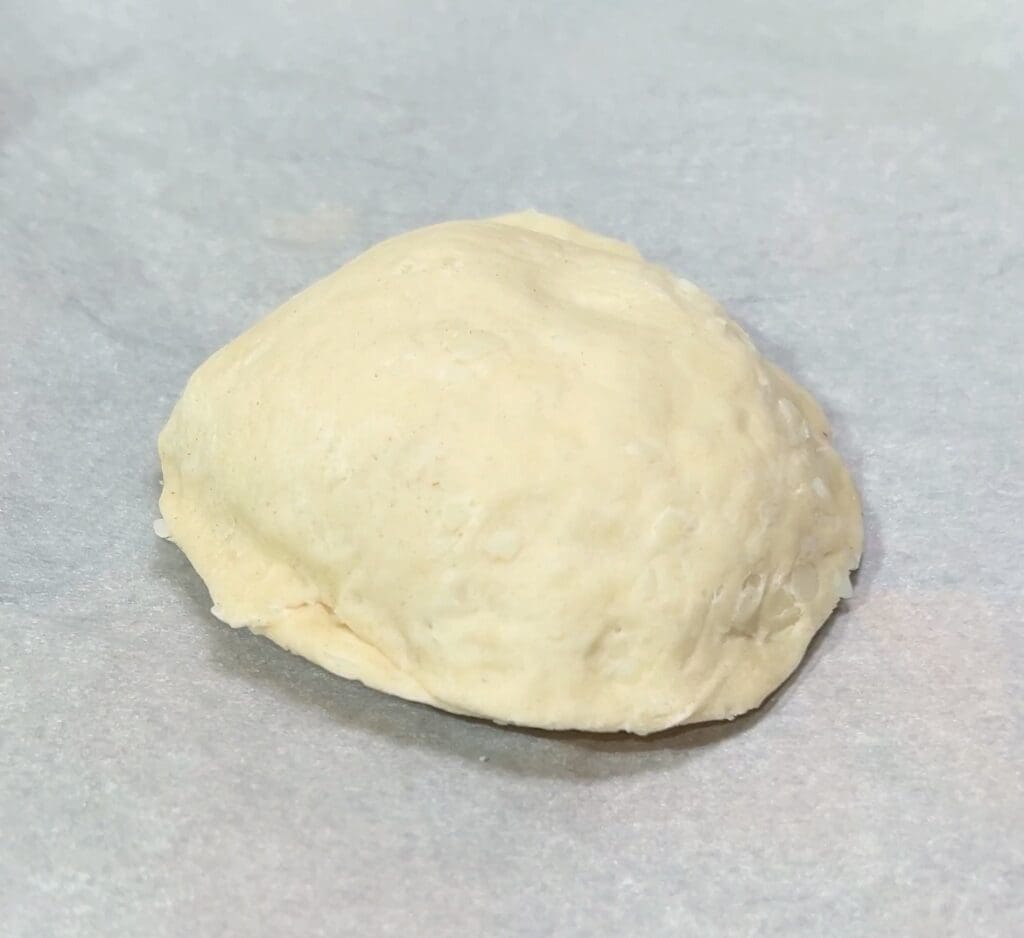 A raw ball of biscuit dough lies on a parchment sheet, showcasing its smooth, lightly floured surface and sealed edges.
