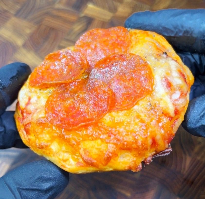 Gloved hands holding a small, round pepperoni stuffed pizza biscuit with melted cheese and crispy edges. The backdrop features a wooden surface.