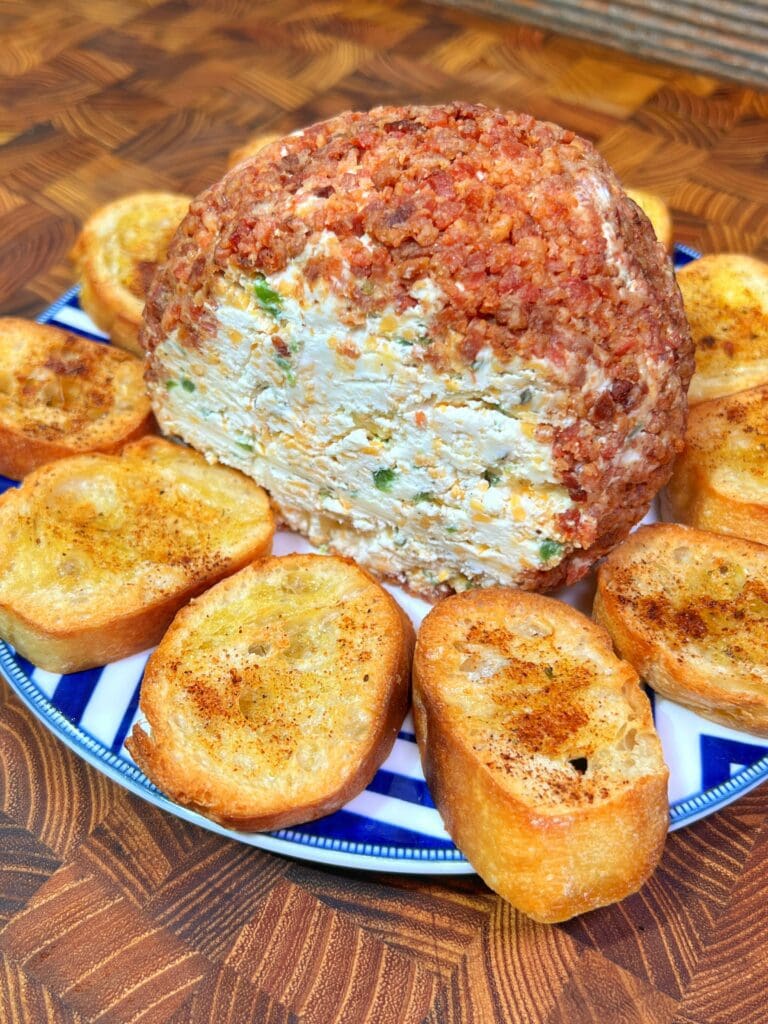 A jalapeno popper cheese ball with a crispy topping on a blue and white plate surrounded by toasted crostini.