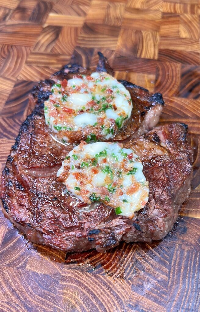 A grilled steak topped with two rounds of blue cheese butter, garnished with chives and bits of bacon, served on a wooden cutting board with a distinct grain pattern.