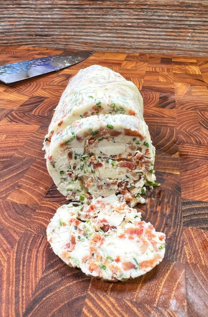 A log of blue cheese compound butter rolled with herbs and pieces of bacon is displayed on a wooden cutting board. Several sliced pieces are placed in front of the log, showcasing the bacon bits and herbs embedded within the cheese.