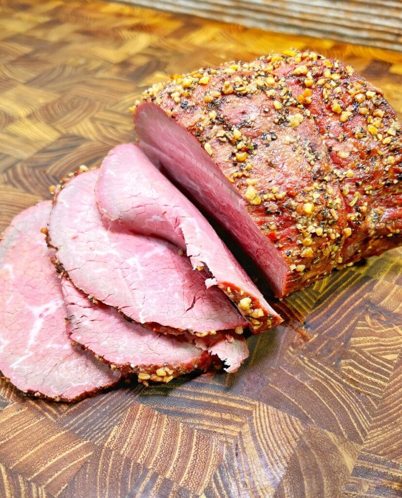 Sliced deli roast beef on a wooden cutting board, seasoned with crushed spices and peppercorns, showcasing its pink interior and textured outer layer.