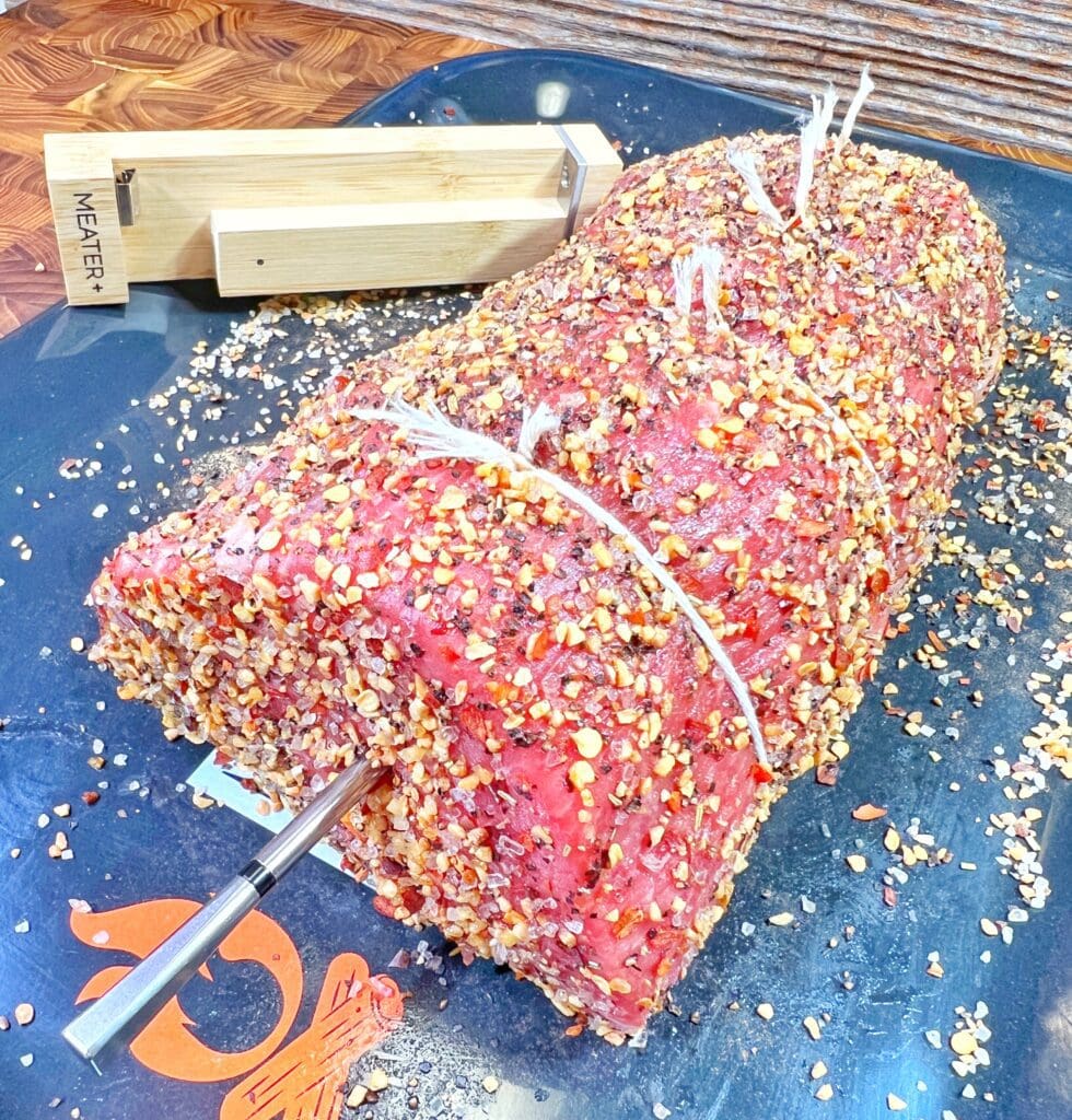 A large, seasoned deli beef roast covered in crushed peppers and spices rests on a black cutting surface. It is tied with white string and has a meat thermometer inserted into it. A wooden box labeled MEATER is seen in the background.