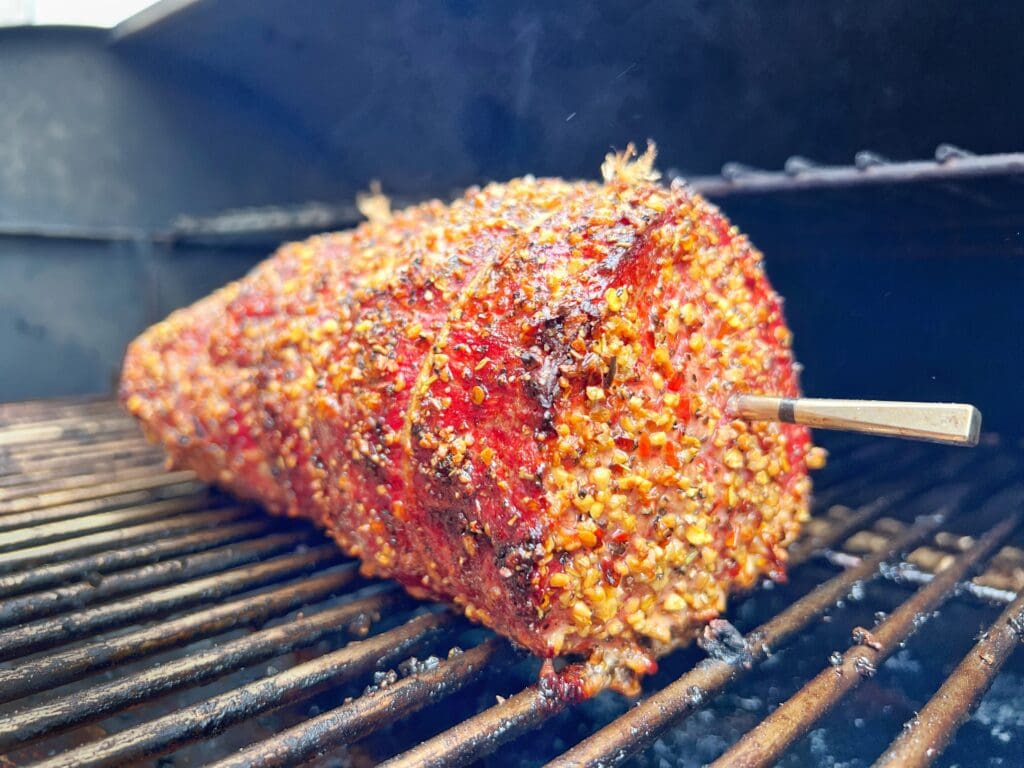 A large piece of well-seasoned deli beef meat is cooking on a grill. The meat is covered in a crust of spices and has a thermometer inserted in one side. The grill grates are visible, and smoke lightly rises around the meat.