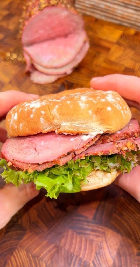 Close-up of hands holding a sandwich of deli roast beef with layers of roast beef, lettuce, and sauce in a soft bun. A sliced roast beef joint is visible in the background on a wooden surface.