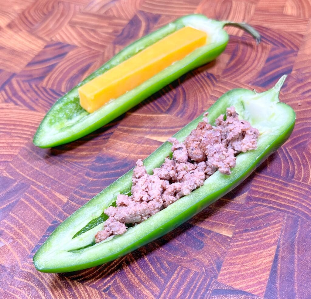 Two halved jalapeños on a wooden surface, one filled with a stick of cheddar cheese and the other with ground meat.