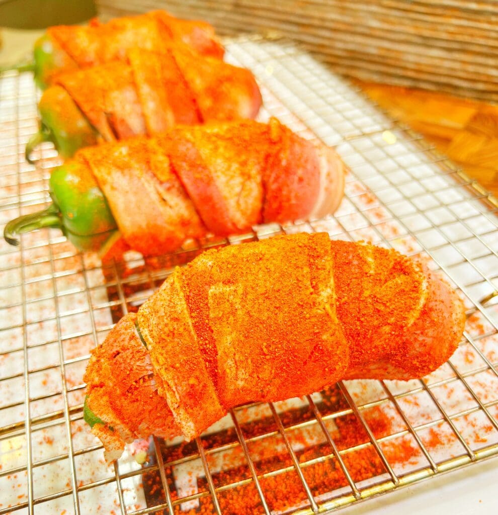 Four hamburger meat stuffed jalapeños wrapped in bacon and coated with seasoning are placed on a wire rack.