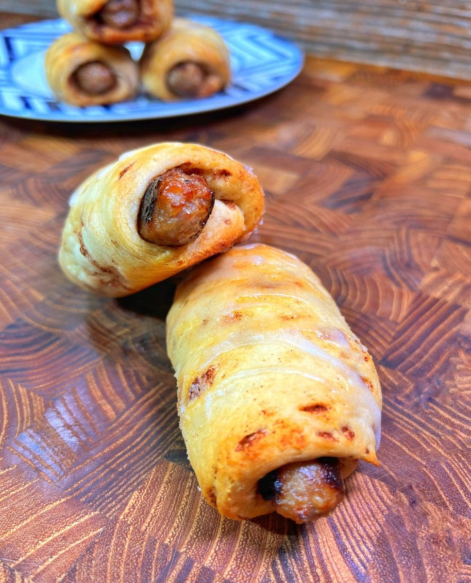 Two sausage rolls made with golden-brown pastry sit on a wooden board. In the background, more sausage rolls are visible on a blue-patterned plate. The surface features a textured pattern.