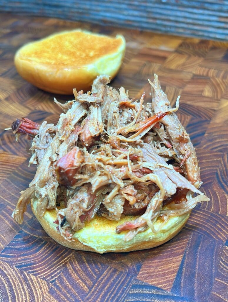 A sandwich with Dr. Pepper pulled pork on a toasted bun is displayed on a wooden surface. The top half of the bun is placed in the background, showing the texture of the shredded pork clearly.
