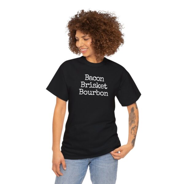 A curly-haired individual wearing the Bacon Brisket Bourbon black T-shirt with white text stands against a plain white background. A tattoo is visible on their right forearm, and their slight smile suggests they're savoring a perfect moment as they gaze down thoughtfully.