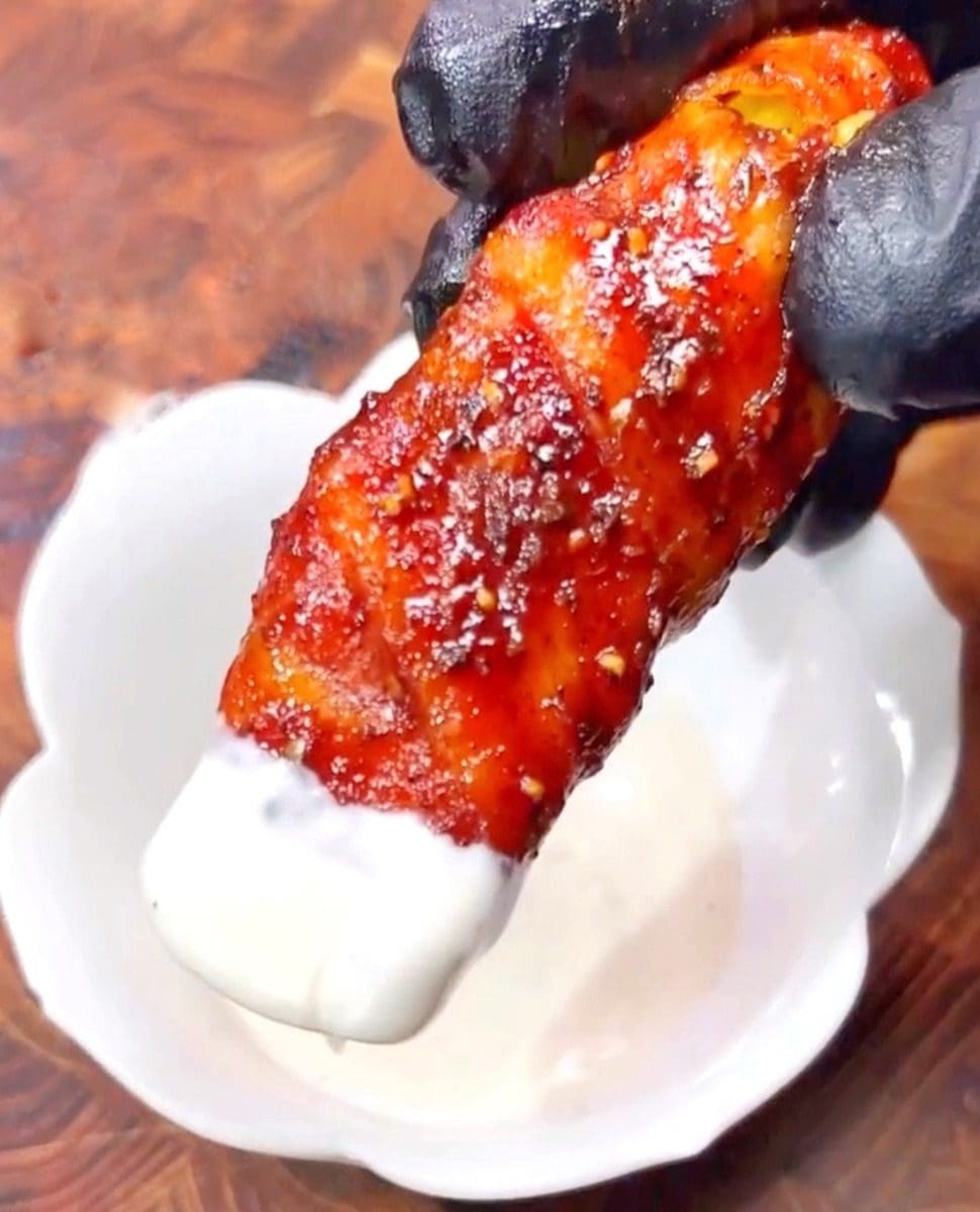 A gloved hand holds a bacon-wrapped chicken tender coated in sauce and sprinkled with seasoning, reminiscent of tantalizing Bacon Cheeseburger flavors. The tender is being dipped into a white creamy sauce in a small white bowl. The background is a wooden surface.
