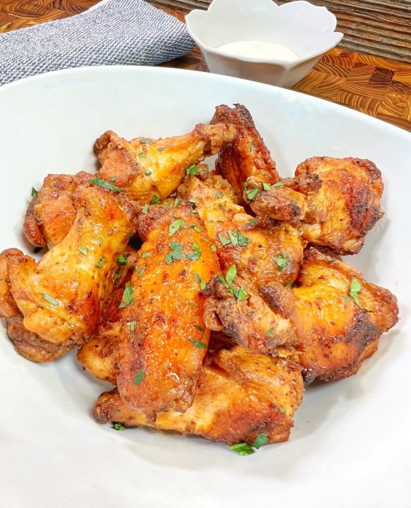 A white bowl filled with crispy, golden-brown chicken wings made following Gordon Ramsay's recipe and garnished with chopped herbs—a perfect, tasty delight. In the background, there's an easy creamy dipping sauce in a small white bowl. A textured cloth napkin is partially visible beside it.