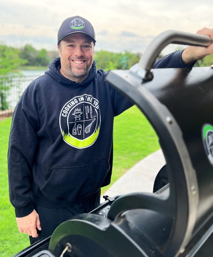 A man wearing a black hoodie and cap, both featuring a Cooking in the Yard logo, smiles while standing next to a barbecue grill in a backyard setting. The grass is bright green, and the sky is overcast.