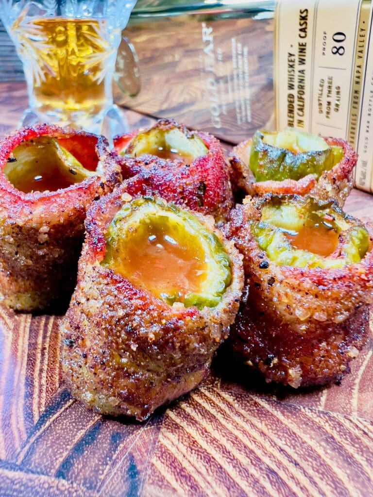 close up image of bacon wrapped bloody mary pickle shots on a wooden cutting board in front of a bottle of whiskey.