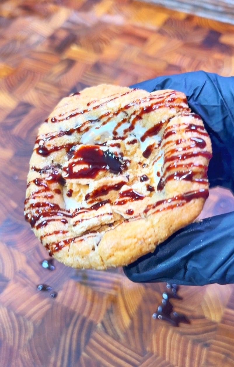 A gloved hand holding a cookie drizzled with chocolate sauce. The cookies surface has a textured appearance, and its held over a wooden cutting board with a geometric pattern.