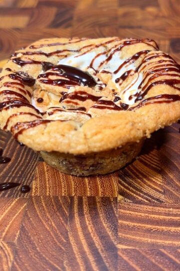 A close-up of a cookie cup filled with gooey marshmallow and drizzled with chocolate sauce, placed on a wooden surface with rich grain patterns.