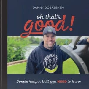 A man wearing a hoodie stands near a grill with a scenic background, smiling. The cover text on the "Oh That's Good! Hard Cover Cookbook" reads "Perfectly Easy Recipes That You Need to Know.