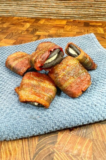 Delicious bacon-wrapped cookies rest on a blue towel atop a wooden surface. The partially cut cookies reveal a creamy filling inside, making them irresistibly tasty.