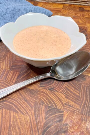 A white scalloped bowl filled with creamy tomato soup rests on a wooden table beside a large silver spoon. A blue cloth is partially visible in the background.