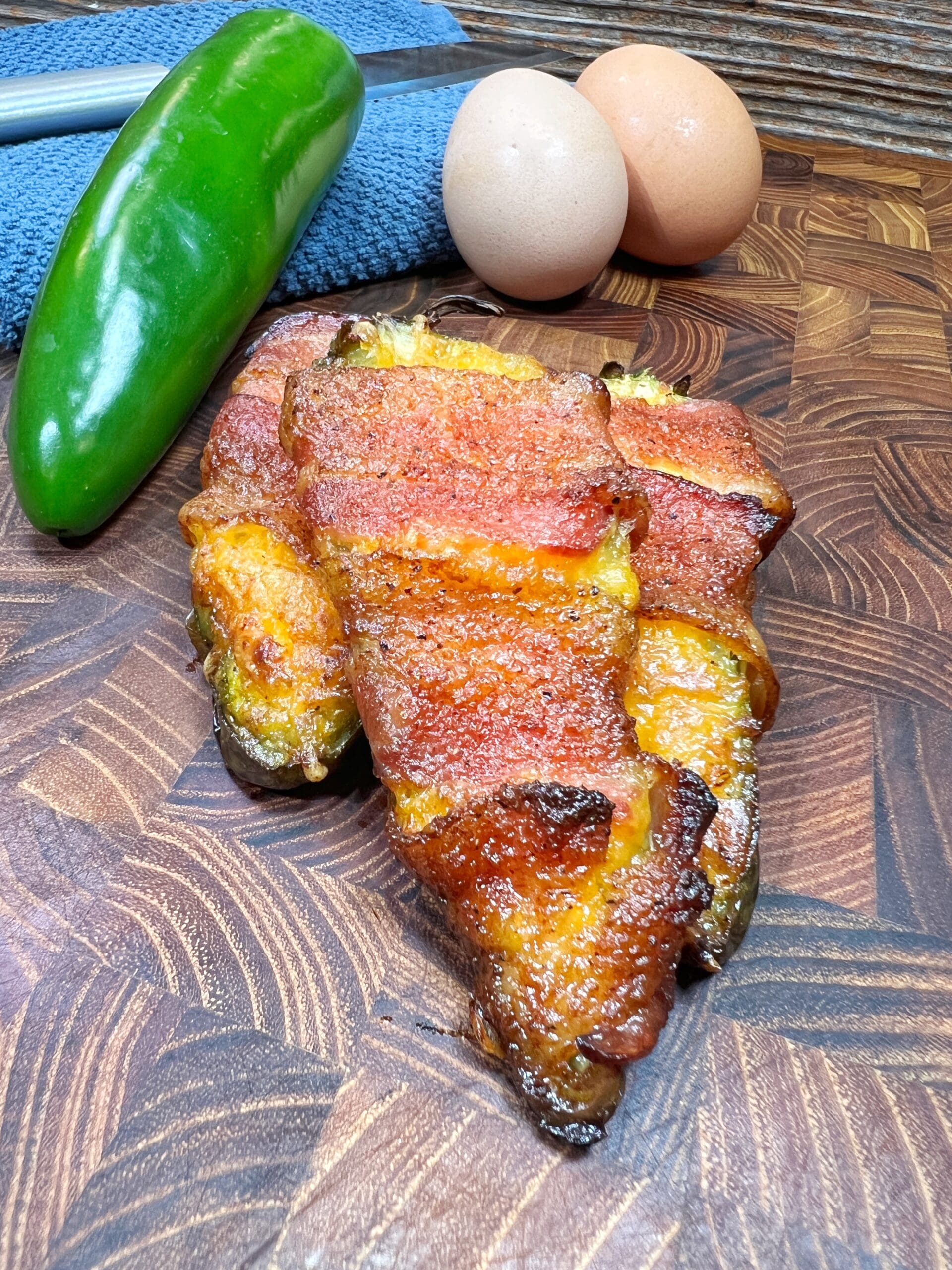 Crispy bacon-wrapped jalapeño poppers on a wooden cutting board, with a whole green jalapeño, two eggs, and a blue towel in the background.