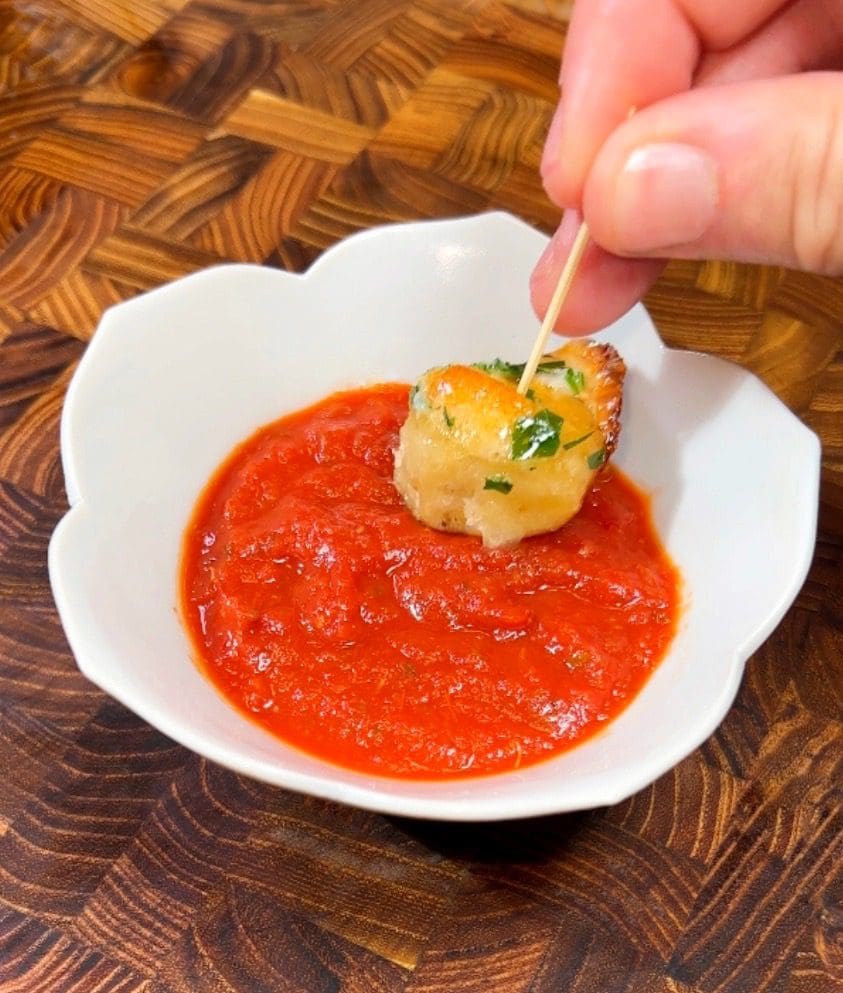 A hand holding a toothpick with a small, breaded appetizer. The appetizer is being dipped into a red tomato sauce in a white bowl, set on a wooden table with a carved pattern.