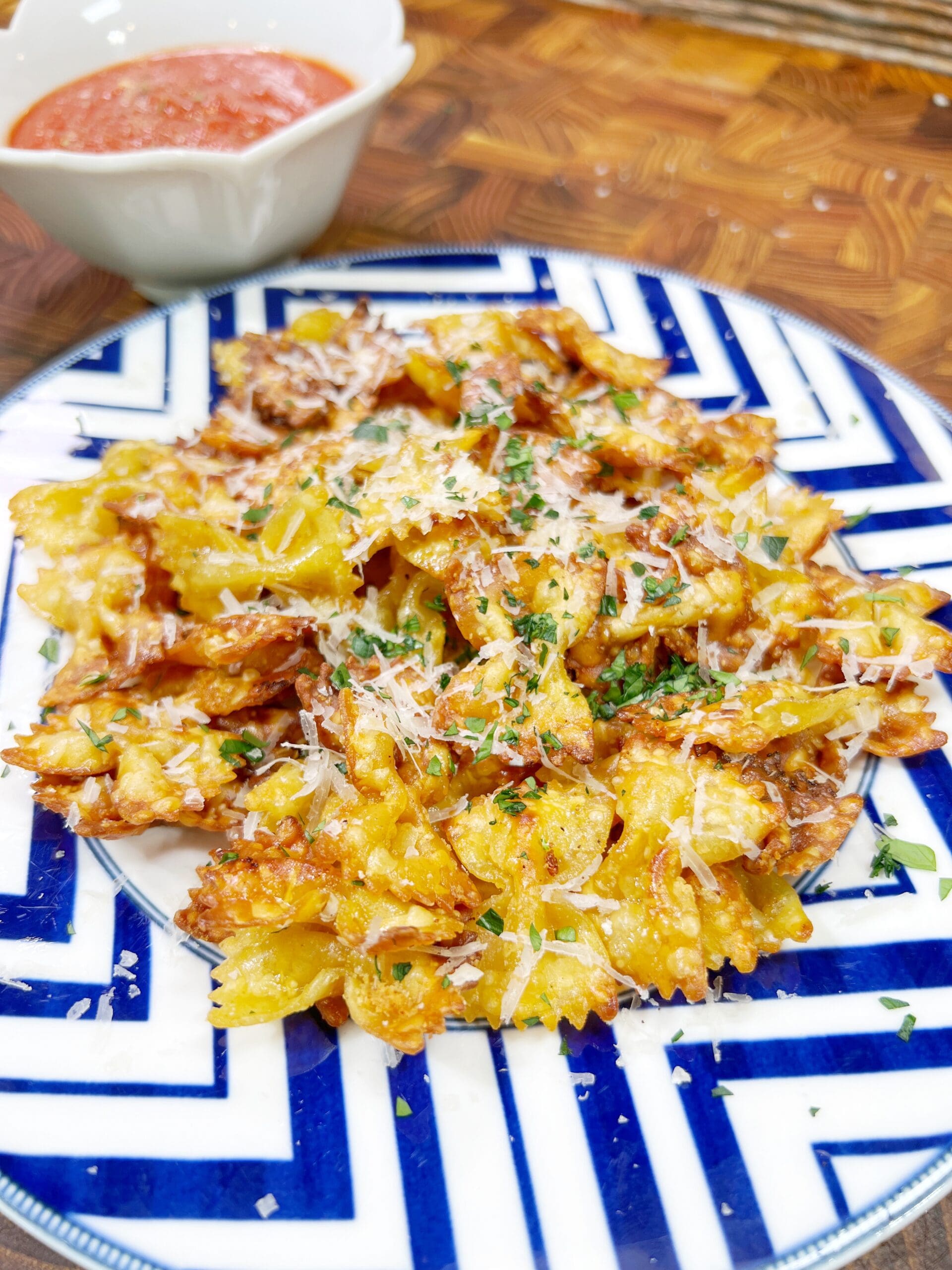 A plate of crispy bow-tie pasta chips garnished with grated cheese and herbs is served on a blue and white patterned plate. A small dish of red dipping sauce is in the background.
