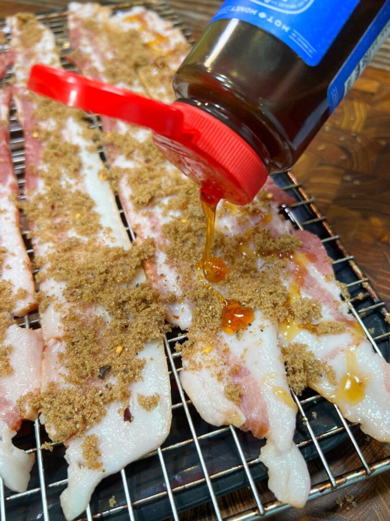 A bottle of syrup is being poured over strips of bacon covered with brown sugar, placed on a wire rack. The rack sits on a wooden surface in preparation of smoked candied bacon.