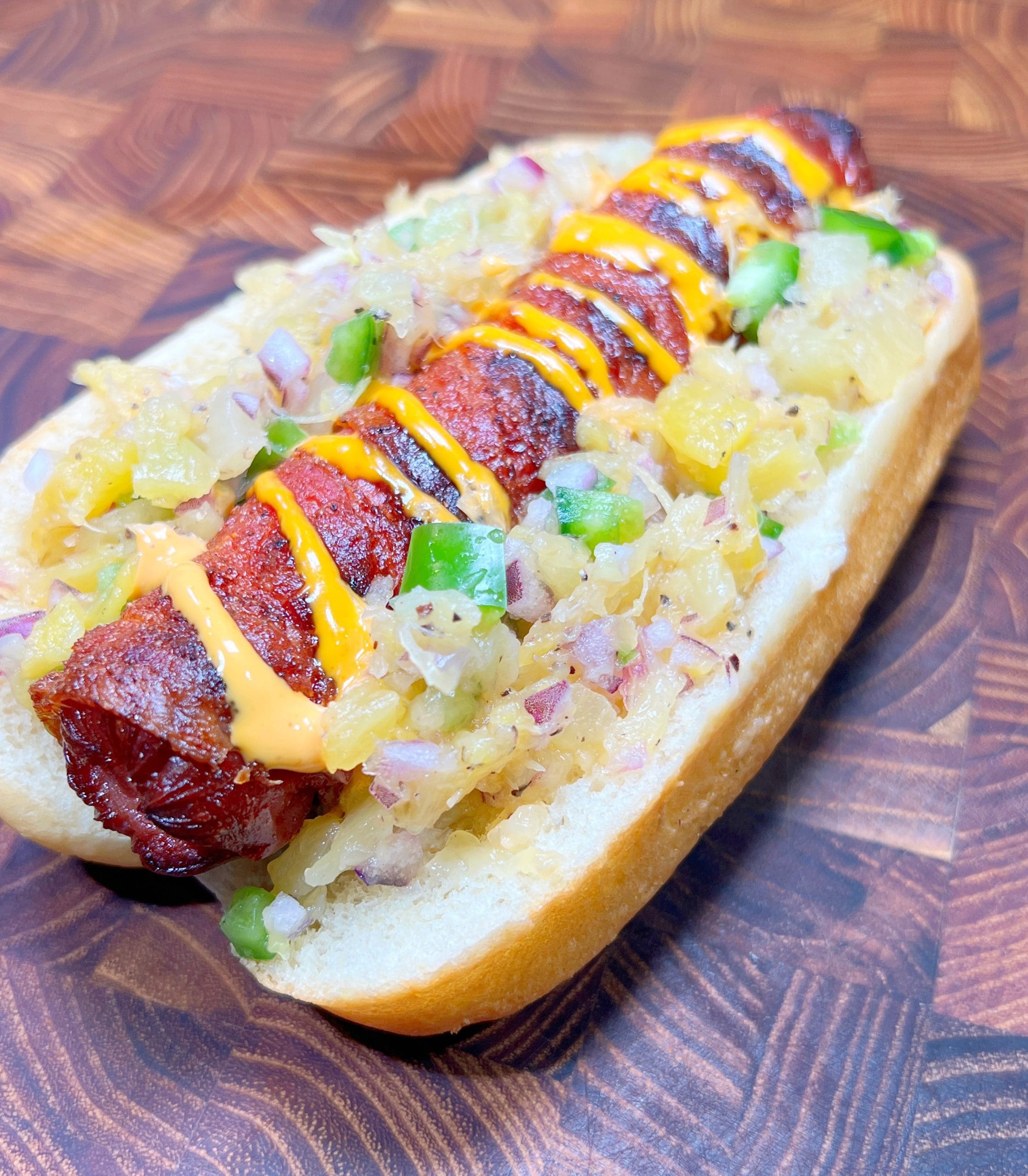A grilled hot dog topped with diced onions, pineapple, jalapeños, and a drizzle of orange sauce, served on a toasted bun. The background is a wooden surface with a rich brown pattern.