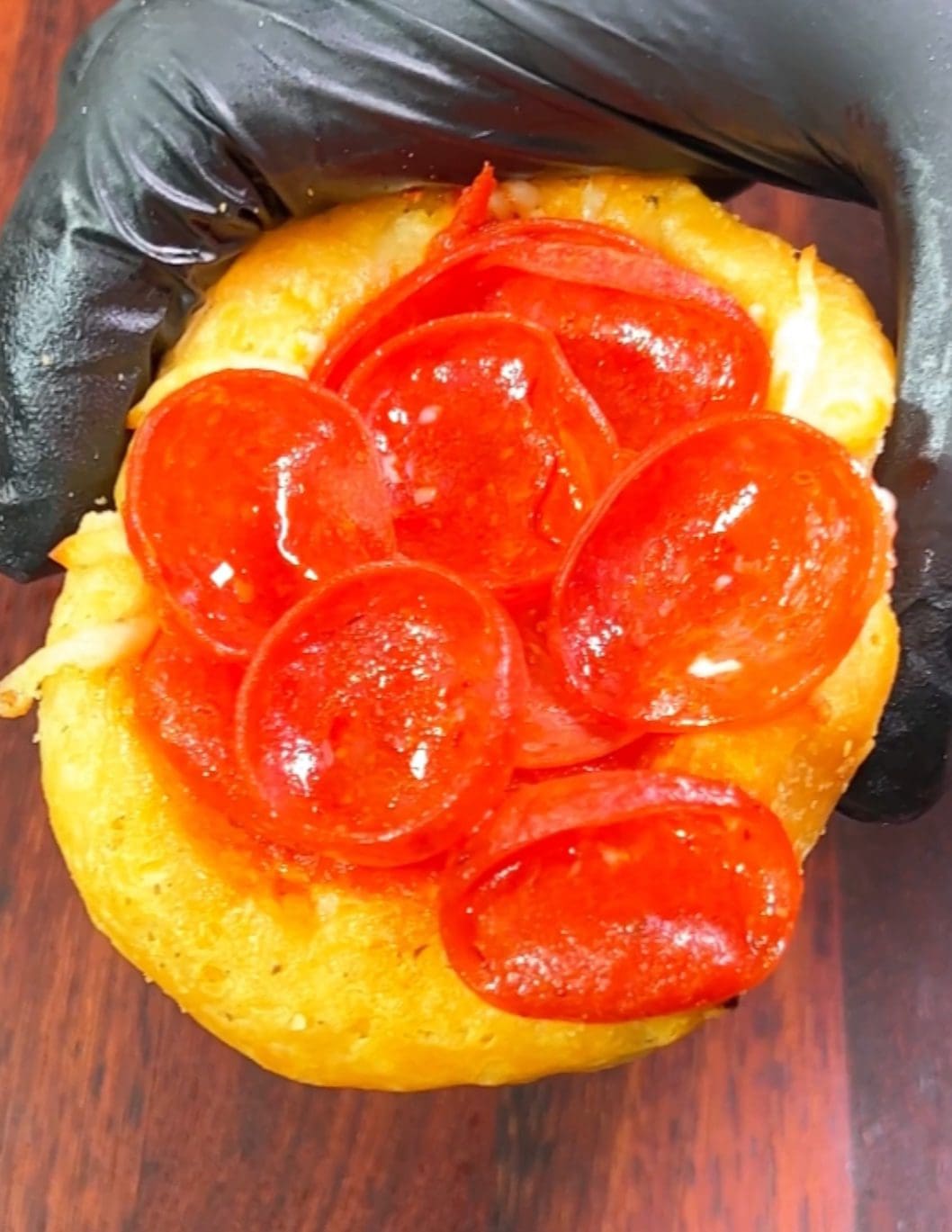 A gloved hand holds a mini pizza made on a small, thick dough base, topped with melted cheese and several slices of shiny pepperoni. The pizza is set against a wooden background.