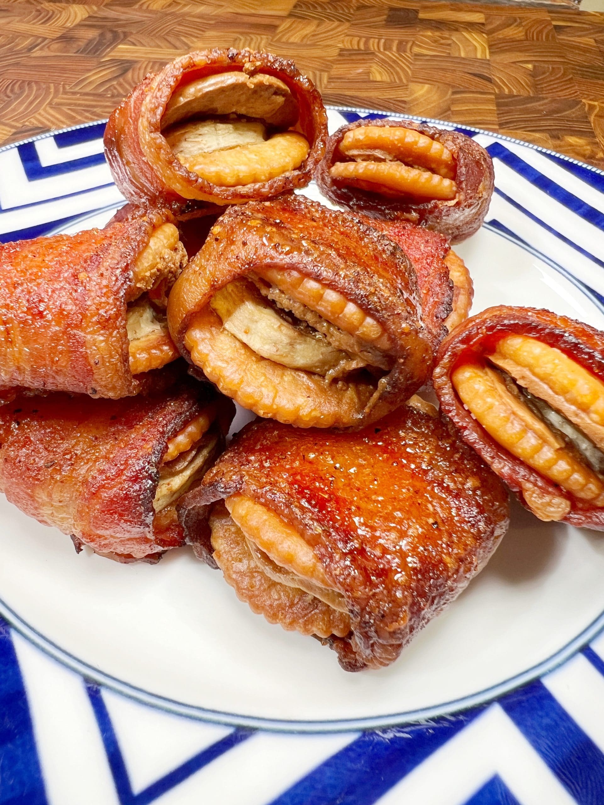 A plate of crispy fried bacon-wrapped treats, each containing a filling resembling crackers or cookies, placed on a white plate with a blue geometric rim design. The background is a wooden surface.