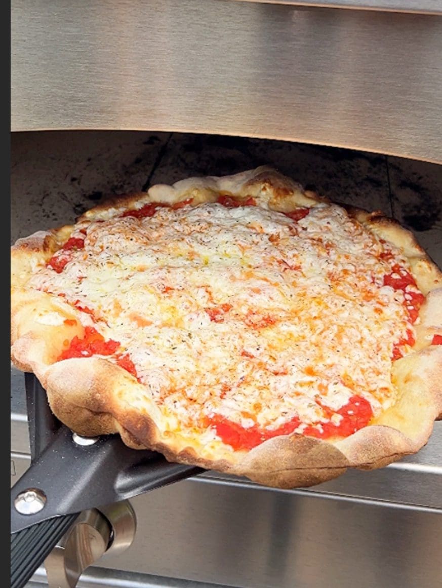 A freshly baked pizza with a crispy crust and melted cheese is being taken out of a pizza oven using a spatula. The pizza has a golden-brown crust and is topped with tomato sauce and cheese.