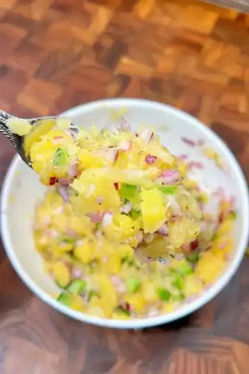 A spoon brimming with pineapple jalapeno relish over a bowl of relish in a white bowl on a wooden surface.