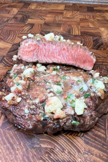 A delicious cooked steak with a slice cut to reveal a juicy pink center, topped with garlic and herbs, displayed on an easy-to-clean wooden cutting board with a checkerboard pattern.
