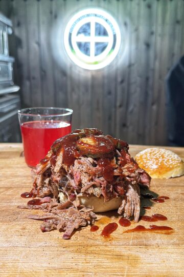 A pulled pork sandwich made from a classic pulled pork recipe is topped with barbecue sauce and pickles, sitting on a wooden surface beside a sesame bun half and a glass of red drink. A circular window is visible in the wooden wall behind.