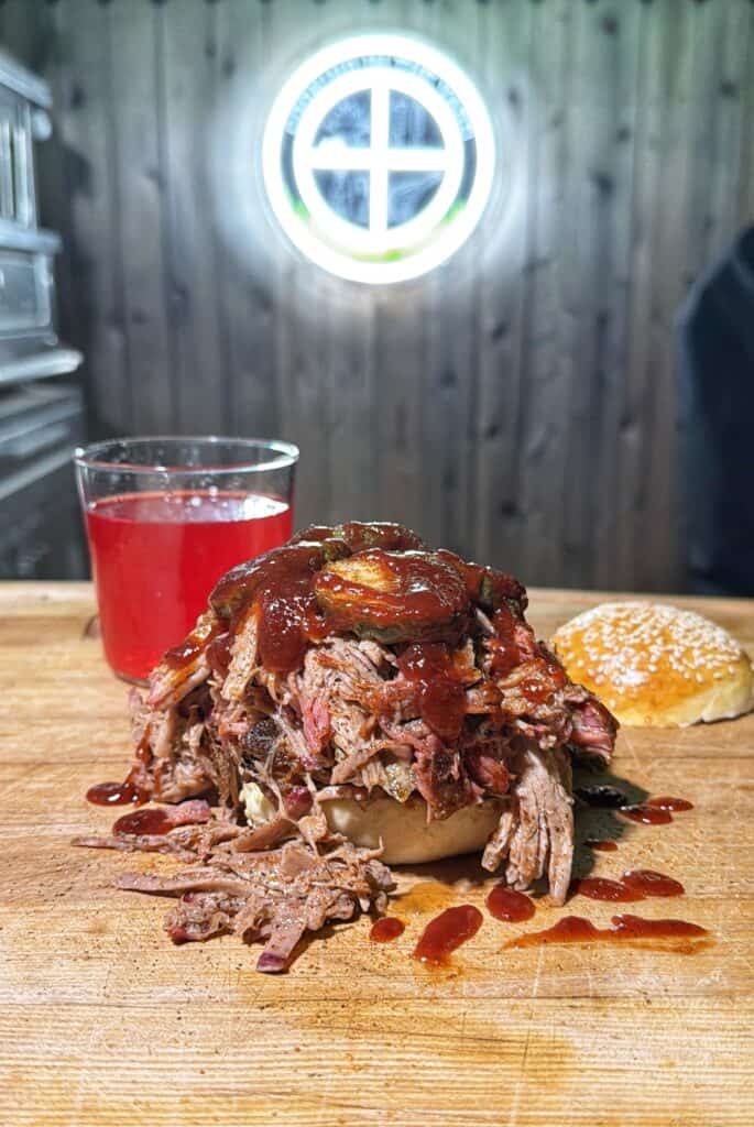 A pulled pork sandwich made from a classic pulled pork recipe is topped with barbecue sauce and pickles, sitting on a wooden surface beside a sesame bun half and a glass of red drink. A circular window is visible in the wooden wall behind.