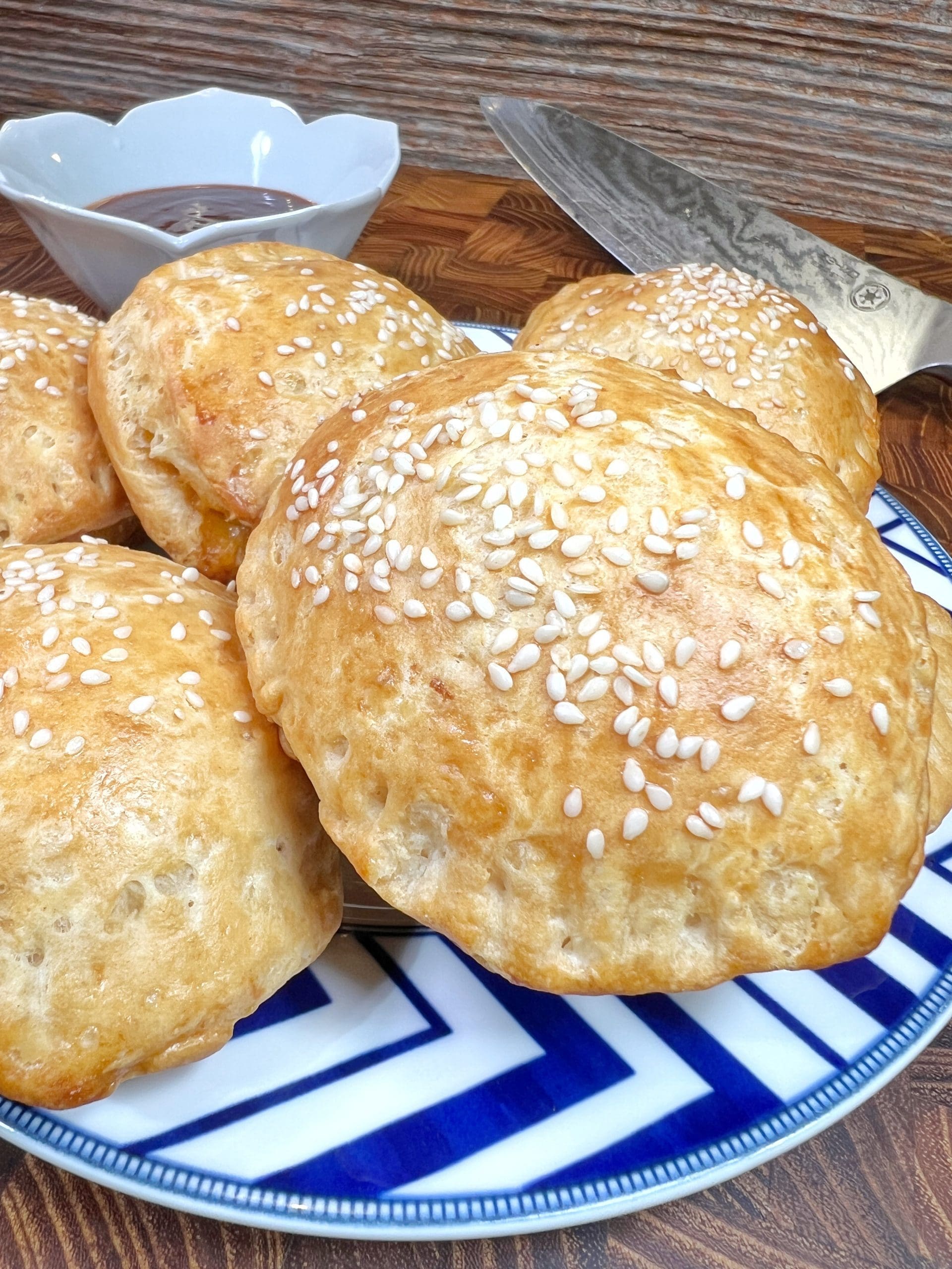 A plate of golden, freshly baked buns topped with sesame seeds. The buns are arranged in a pile on a decorative blue and white plate. A dipping sauce and a knife are visible in the background on a wooden surface.