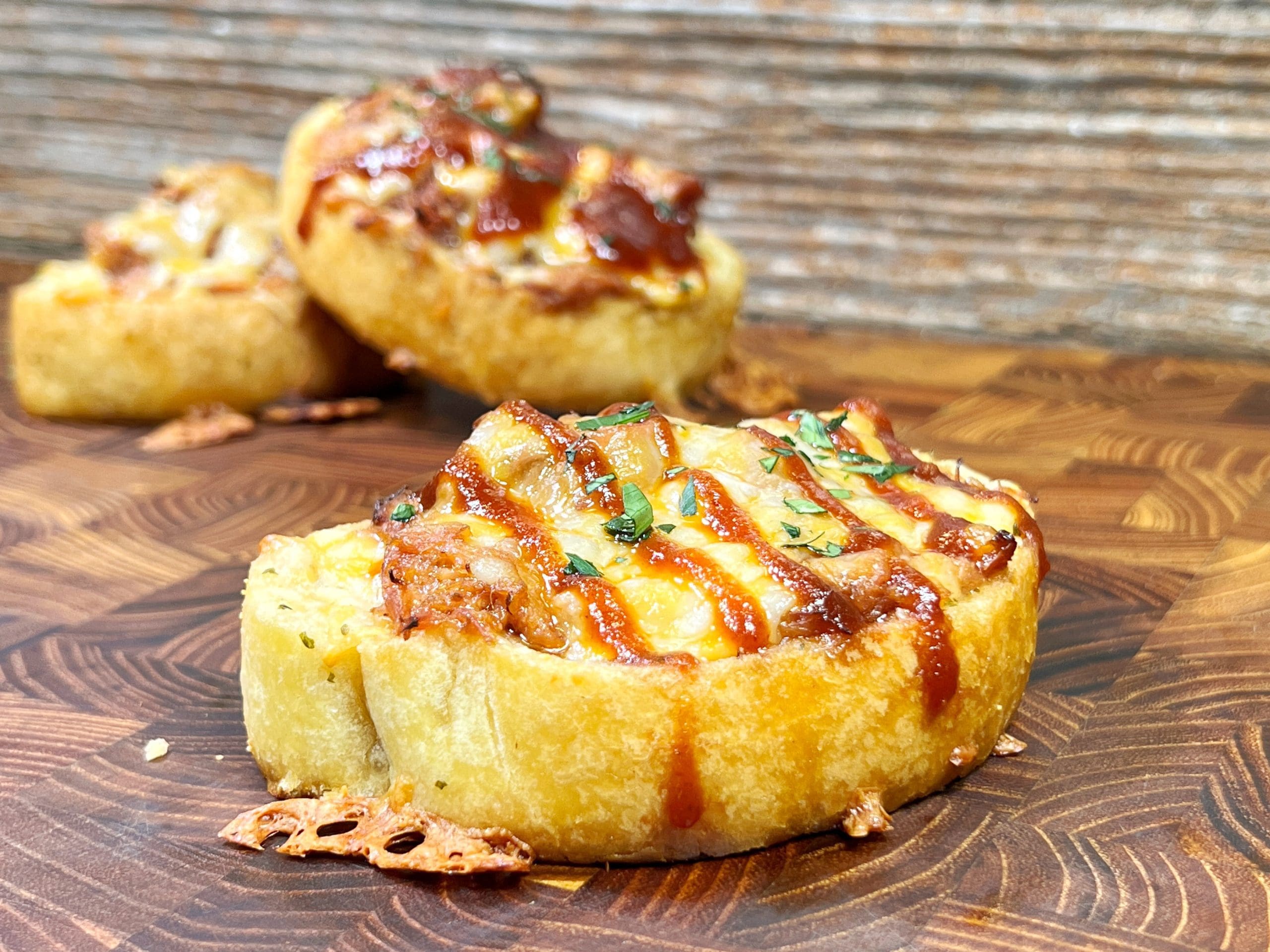 Slices of toasted bread topped with melted cheese, chopped herbs, and a drizzle of barbecue sauce are displayed on a wooden surface. The bread appears crispy and is garnished with cooked meat pieces.