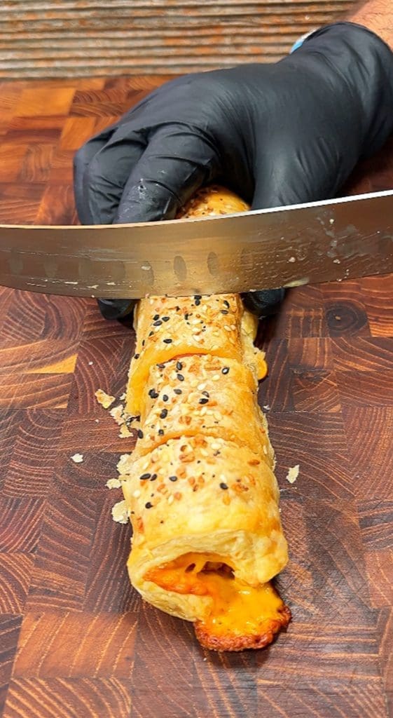A gloved hand uses a large knife to slice a golden-brown pastry filled with melted cheese and hotdog. Topped with sesame seeds and seasoning, the pig in a blanket is sitting on a wooden cutting board.