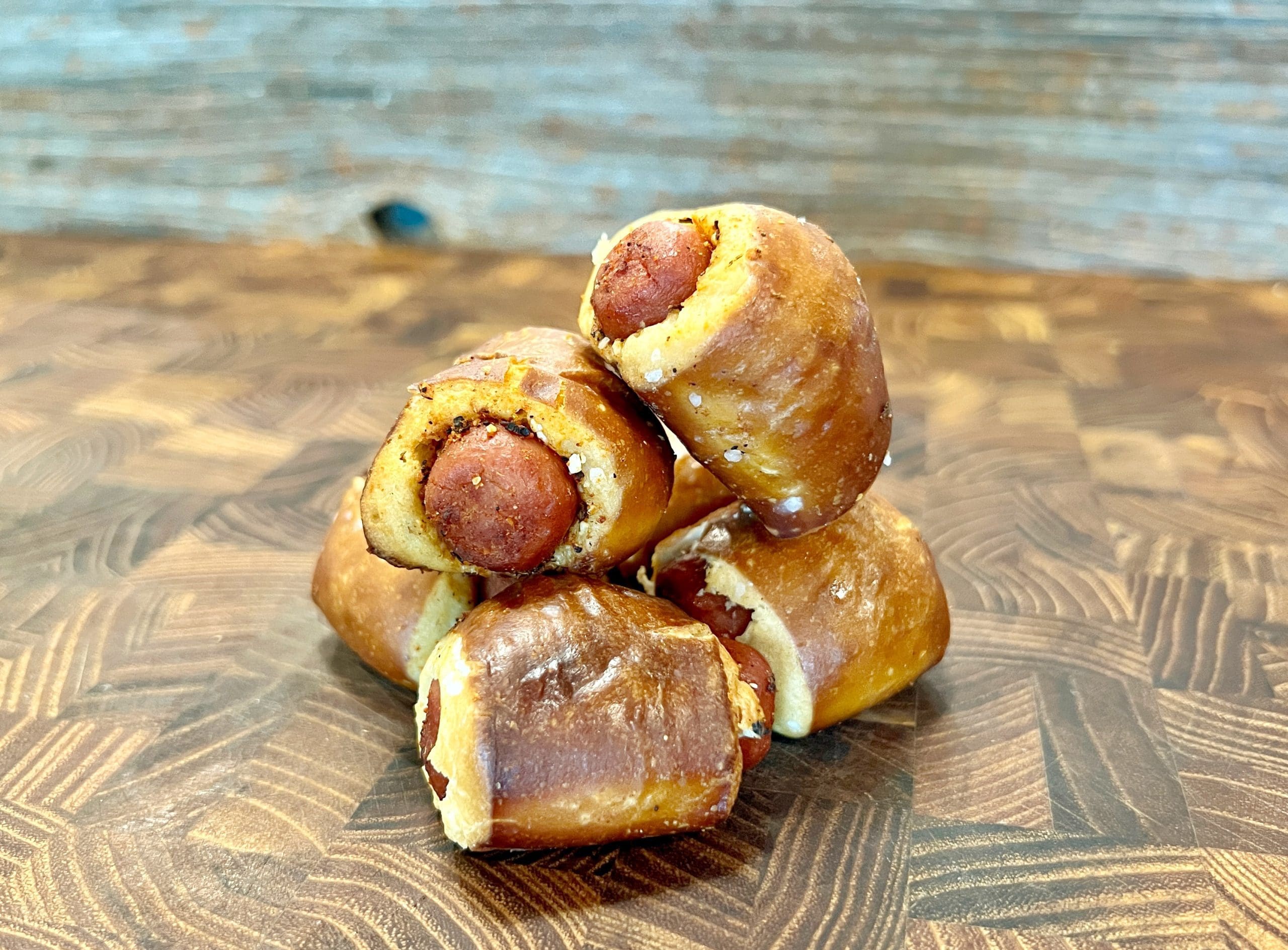 A stack of pretzel-wrapped mini sausages is arranged on a wooden surface. The pretzels are golden brown with a sprinkle of salt on top, creating a savory and appetizing appearance.