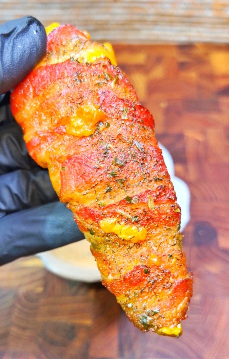 A gloved hand holding a bacon-wrapped jalapeño stuffed with melted cheese. The jalapeño appears roasted and seasoned, with a blurred wooden surface in the background.