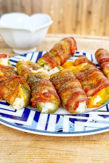 A plate of bacon-wrapped jalapeño pickle poppers filled with cream cheese sits on a wooden surface, with a white bowl in the background. The poppers are golden and slightly browned.